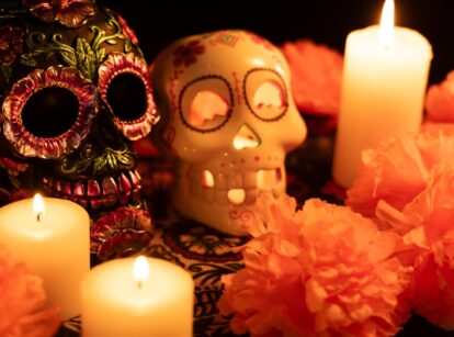 Two Día de los Muertos skulls on a tablecloth: one decorated with metal flowers, and the other with a lit candle and surrounded by bright orange marigold flowers.