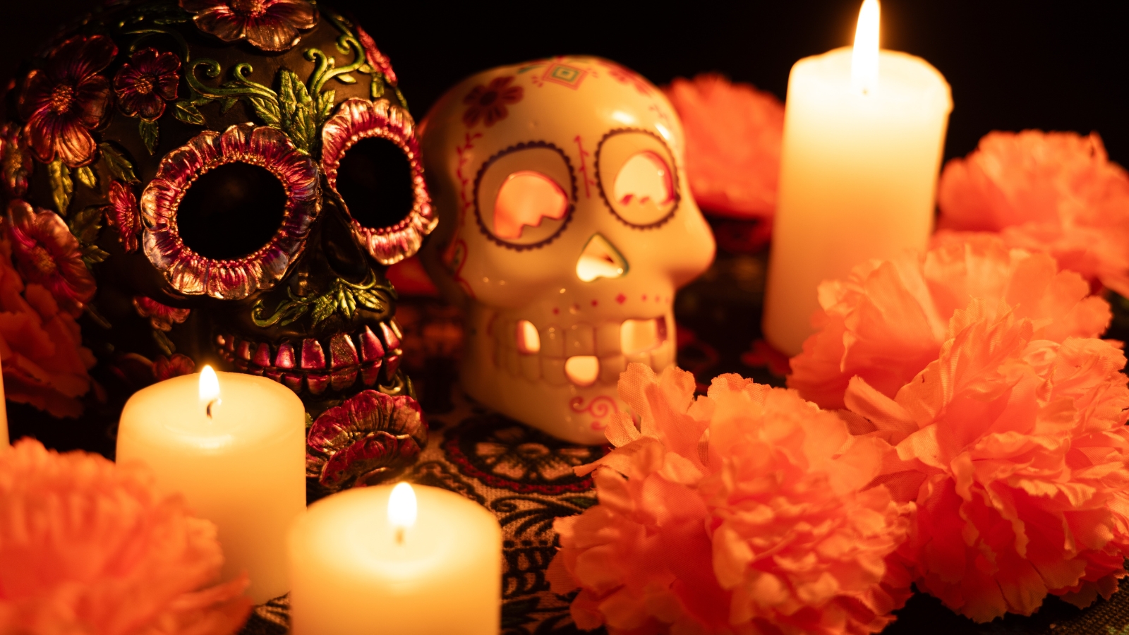 Two Día de los Muertos skulls on a tablecloth: one decorated with metal flowers, and the other with a lit candle and surrounded by bright orange marigold flowers.