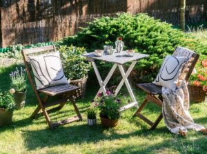 Two garden chairs and a wooden table on a grassy patio, surrounded by potted plants, with a bushy fir bush in the background.