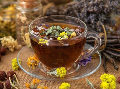 Close up of glass cup filled with freshly brewed herbal tea among various dried herbs and flowers.