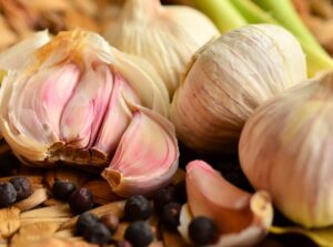 Close-up of garlic heads with cloves covered in white and pinkish papery skins, arranged in a wicker basket with scattered black peppercorns.