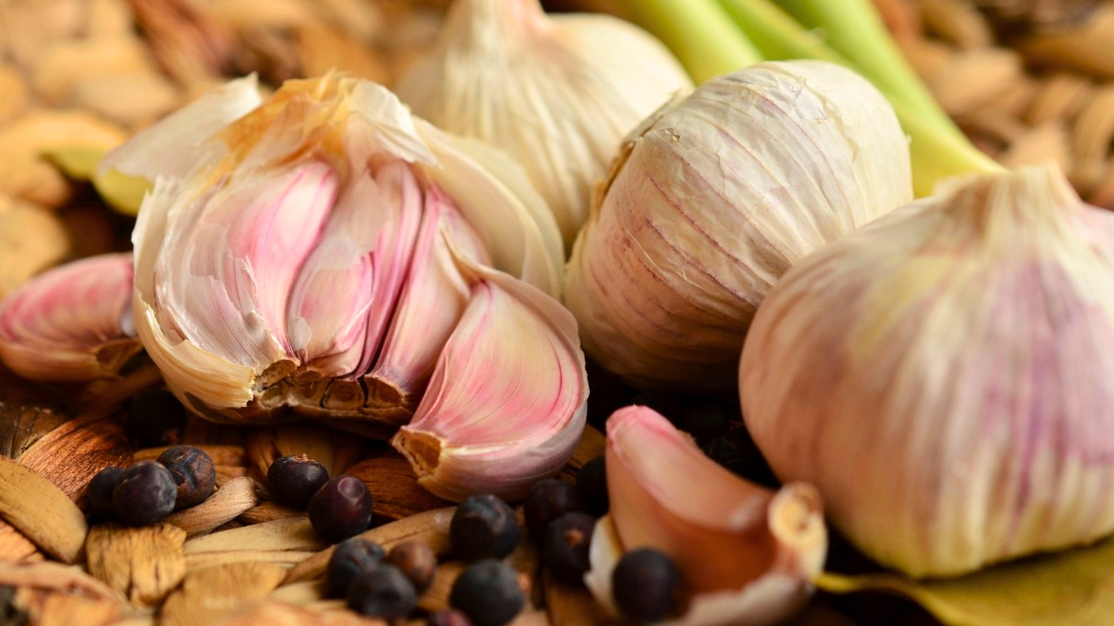 Close-up of garlic heads with cloves covered in white and pinkish papery skins, arranged in a wicker basket with scattered black peppercorns.