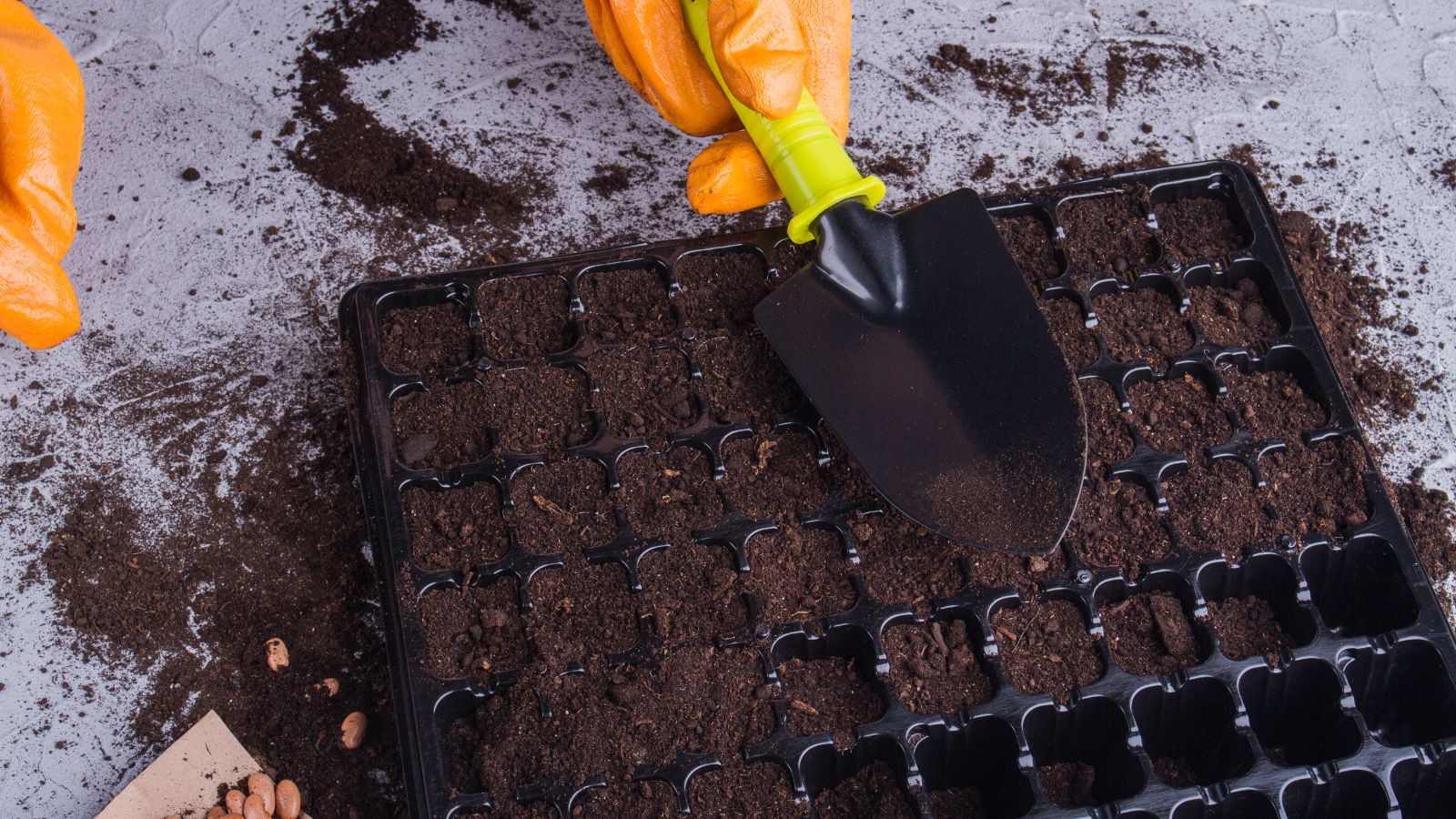 A shot of a tray that is being filled  with soil by a person who is using a small garden shovel with yellow handles and is wearing orange gloves, all placed on a white surface in a well lit area 