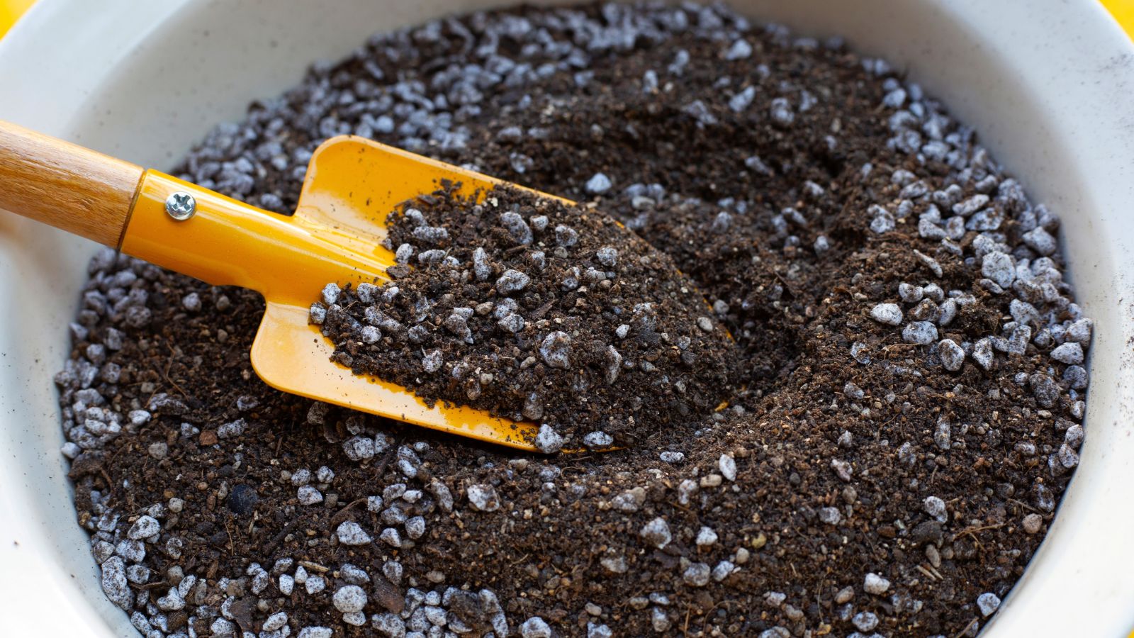 An overhead shot of a white container filled with potting mix and a small yellow shovel in a well lit area